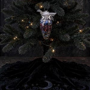 May include: A clear, teardrop-shaped glass ornament filled with small, multi-colored polished stones, topped with a silver pentacle and triple moon charm and a white ribbon, hanging from a dark green Christmas tree above a black velvet tree skirt.