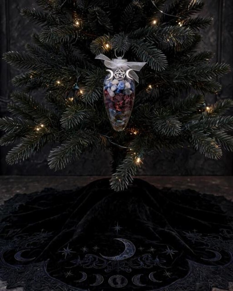 May include: A clear, teardrop-shaped glass ornament filled with small, multi-colored polished stones, topped with a silver pentacle and triple moon charm and a white ribbon, hanging from a dark green Christmas tree above a black velvet tree skirt.