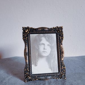May include: Ornate bronze-toned picture frame with a portrait of a woman. The frame features a detailed floral design around the edges. The portrait is in black and white, adding a vintage aesthetic. The frame is rectangular and stands upright.