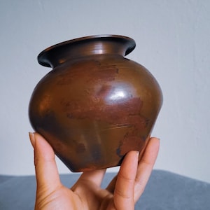 May include: A small, antique copper pot with a rounded body and a flared rim. The pot has a dark, aged patina with visible variations in color. The pot is held in a hand against a neutral background.