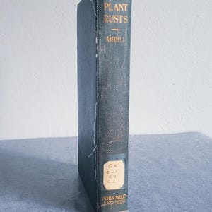 May include: A vintage book titled "Plant Rusts" by Arthur, with a teal cover and gold lettering. The book has a worn appearance, with a label on the spine. The book is standing upright on a gray surface.