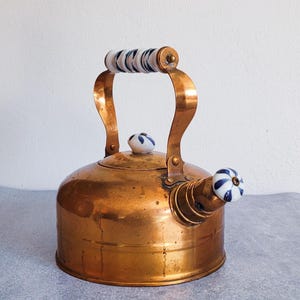 May include: A copper-colored tea kettle with a curved handle and spout. The handle and spout knob are white with blue and black striped and floral designs. The kettle has a round body and a lid with a small knob.