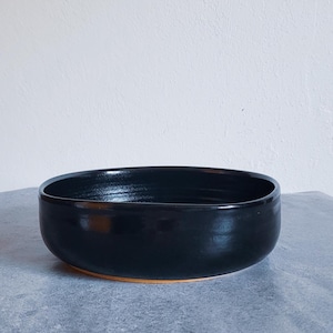 May include: A round, shallow bowl with a smooth, glossy black interior and exterior finish. The bowl features a low profile with slightly curved sides and a visible terracotta-colored base rim, showcasing a simple, functional design.