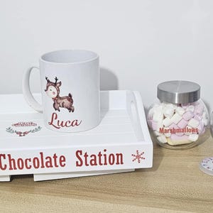 May include: A white wooden tray with cut-out handles, labelled "Luca's Hot Chocolate Station" in red text, holds a mug with a deer graphic and the name "Luca." Beside the tray are glass jars filled with marshmallows, chocolates, and hot chocolate mix.