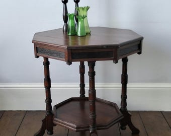 May include: An octagonal wooden side table with a dark brown finish, featuring a lower shelf and four turned legs. On the tabletop sit two tall, thin, dark brown taper candles and two small, translucent, light green glass vases with ruffled rims.
