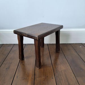 May include: A small, rectangular wooden stool with a dark brown finish and visible wood grain. The stool features four straight, sturdy legs and a flat top surface, showcasing a simple, functional design with signs of wear on the edges.