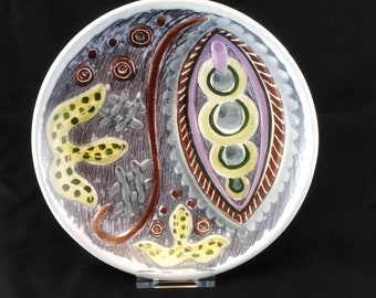 May include: A white ceramic plate with a colorful abstract design. The design features yellow, brown, purple, and gray shapes and lines.