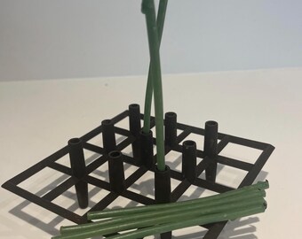 May include: A black metal grid with multiple vertical tubes, used as a flower vase. Green stems are inserted into the tubes, with some stems laying on the grid.