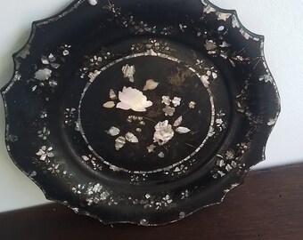 May include: A black circular tray with a scalloped edge, decorated with a floral design made of mother of pearl. The tray has a raised rim and a smooth surface.