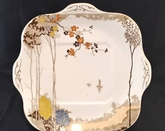 May include: A white square serving plate with a scalloped edge and a delicate floral design in shades of brown, orange, and gold. The design features a tree branch with blossoms and a landscape scene.