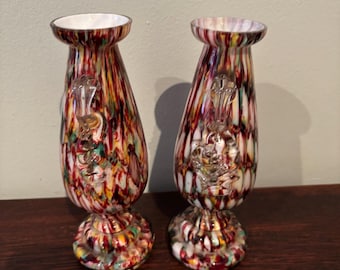 May include: Two decorative glass vases with a colorful, speckled design in red, white, yellow, and green. Each vase has a clear, ornate glass embellishment and a white interior. The vases are displayed on a dark wooden surface.