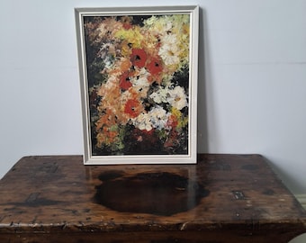May include: An abstract floral painting in a white frame. The artwork features a vibrant mix of colors, including red, yellow, white, and green, creating a textured, impressionistic effect. The painting is displayed on a rustic, dark wooden table.