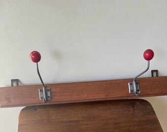 May include: A wooden coat rack with two metal hooks. Each hook has a red ball at the end. The rack is mounted on a wall. The hooks are made of metal and are attached to a wooden board.
