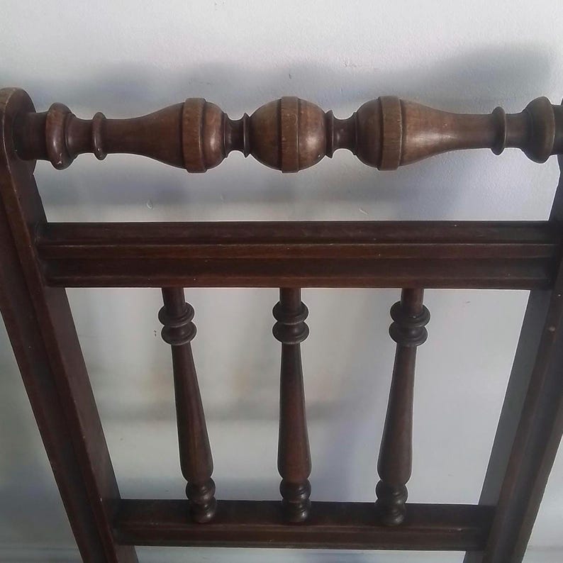 Puede incluir: Un respaldo de silla de madera con un riel superior decorativo y cuatro husos verticales. El respaldo de la silla es de color marr&oacute;n oscuro y tiene un acabado liso.