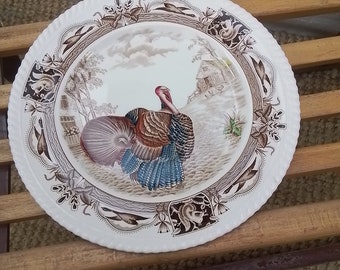 May include: A white ceramic plate with a brown and blue turkey design. The plate has a decorative border with floral and leaf motifs. The turkey is standing in a field with a small house in the background.