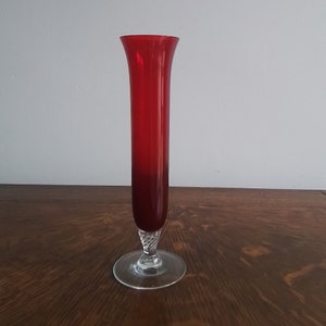 May include: A tall, red glass vase with a flared top and a clear, circular base. The vase has a slender, cylindrical body with a twisted design near the base. The vase is approximately 30 cm tall.
