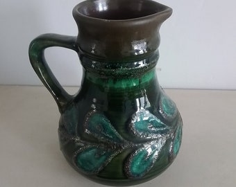 May include: A green ceramic pitcher with a brown rim and a leafy design. The pitcher has a handle and a rounded body.
