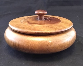 Puede incluir: Una caja redonda de madera con tapa. La caja está hecha de madera marrón claro con una perilla de madera marrón oscuro en la tapa.