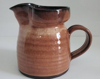 May include: A brown ceramic pitcher with a black rim and handle. The pitcher has a glazed finish and a slightly rounded shape.