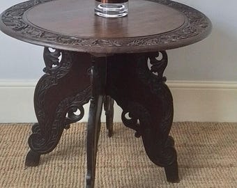 May include: A dark brown wooden table with intricate carvings on the top and legs. The table has a round top and three legs that are shaped like stylized leaves. The table is sitting on a brown rug.