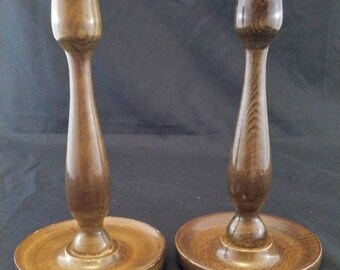 May include: Two wooden candle holders with a brown finish. The candle holders have a round base and a tall, slender stem.
