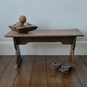 May include: A low wooden bench with a rectangular top and curved legs. On the bench sits a dark wooden bowl containing three spherical objects. A pair of dark-coloured geta sandals with braided straps are positioned on the floor near the bench legs.