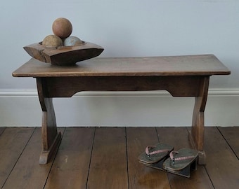 May include: A low wooden bench with a rectangular top and curved legs. On the bench sits a dark wooden bowl containing three spherical objects. A pair of dark-colored geta sandals with braided straps are positioned on the floor near the bench legs.