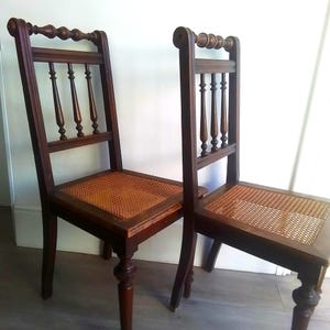 Puede incluir: Dos sillas de madera antiguas con asientos de ca&ntilde;a. Las sillas tienen un acabado marr&oacute;n oscuro y detalles intrincados en el respaldo.