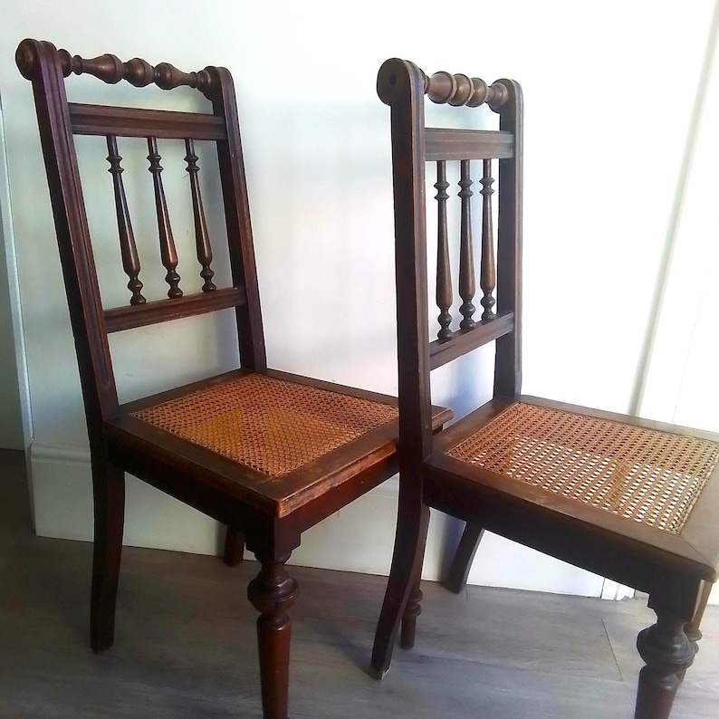 Puede incluir: Dos sillas de madera antiguas con asientos de ca&ntilde;a. Las sillas tienen un acabado marr&oacute;n oscuro y detalles intrincados en el respaldo.