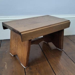 May include: A small wooden step stool. The rectangular top and angled legs are made of light brown wood with a visible grain. The legs have white accents at the base. A simple, functional piece of furniture, suitable for various uses.