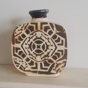 May include: A white ceramic vase with a geometric design in brown. The vase has a square base and a rounded body. The design is a repeating pattern of squares and circles.