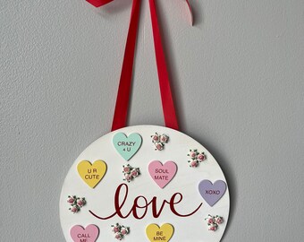 May include: A white round wooden sign with the word "love" written in red cursive. The sign is decorated with colorful candy hearts with various messages, including "UR CUTE", "CRAZY 4 U", "SOUL MATE", "XOXO", "CALL ME", "KISS ME", "BE MINE", and "HUG ME".