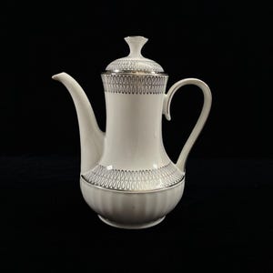 Puede incluir: Una cafetera de porcelana blanca con tapa y asa. La cafetera presenta un patrón decorativo plateado alrededor de la parte superior e inferior. El pico es largo y curvado. El fondo es negro.