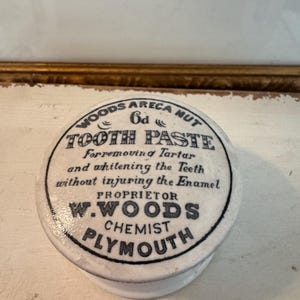 Peut inclure: Un pot en céramique blanc vintage avec une étiquette noire et blanche indiquant "WOODS ARECA NUT TOOTH PASTE". L'étiquette comprend également le texte "PLYMOUTH CHEMIST".