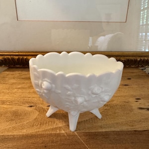 May include: A white, footed bowl with a scalloped rim and embossed floral design. The bowl has three small legs and is made of opaque glass. The bowl is sitting on a wooden surface.