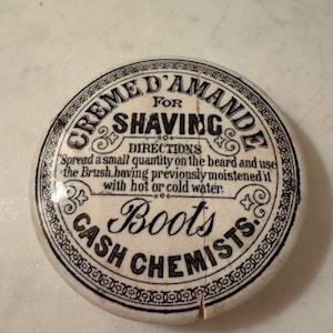 May include: A round, off-white ceramic lid featuring black printed text that reads CREME D'AMANDE FOR SHAVING, DIRECTIONS Spread a small quantity on the beard and use the Brush, having previously moistened it with hot or cold water, and BOOTS CASH CHEMISTS.