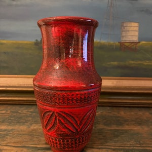 Puede incluir: Un jarrón de cerámica rojo y negro con una superficie texturizada y patrones florales tallados. El jarrón tiene una base ancha y una parte superior ligeramente ensanchada. El fondo presenta una pintura de paisaje con un molino de viento y una torre de agua.