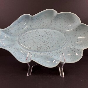 May include: A light blue ceramic serving dish shaped like a leaf with a speckled glaze. The dish is on a clear acrylic stand.