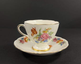 May include: A white teacup and saucer with a floral pattern in shades of pink, orange, yellow, and blue. The cup and saucer have a gold trim.