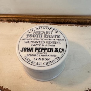 May include: A vintage white ceramic jar of "Cracroft's Areca Nut Tooth Paste." The jar has a circular lid with black text, including "John Pepper & Co, London." The product is described as "Prepared from the Cincalese Recipe."