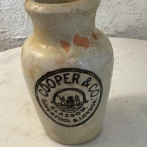May include: A beige stoneware jar with a narrow neck and a wide base. The front features a circular black stamp with the text COOPER & CO LIVERPOOL GLASGOW & LONDON surrounding a central emblem. The surface has light orange discoloration.
