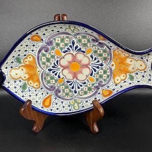 Puede incluir: Un plato decorativo de cerámica con forma de pez y diseño floral pintado a mano. El plato presenta un borde azul y un patrón colorido con acentos naranjas, verdes, morados y rosas. Se exhibe sobre un soporte de madera.
