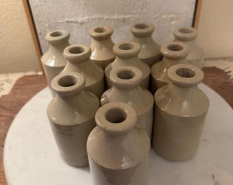 May include: A collection of ten small, cylindrical stoneware bottles with a smooth, light beige glaze. Each bottle features a narrow neck and a flared rim, arranged in a cluster on a circular surface. The bottles have a uniform, simple design.