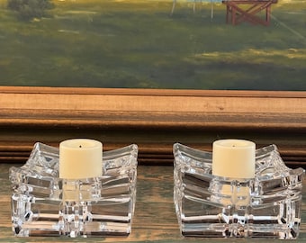May include: Two clear glass candle holders with square bases and a wavy design. Each holder has a white candle inside.