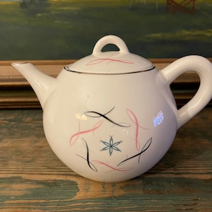 Może przedstawiać: Biały czajnik z różowym i czarnym wzorem kwiatowym. Czajnik ma okrągły korpus i zakrzywiony dzióbek.