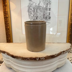 May include: A cylindrical, tan-colored stoneware crock with a slightly flared rim. The antique kitchenware is displayed on a white, scalloped shelf, with a framed architectural print in the background. The crock is approximately 5 inches tall.