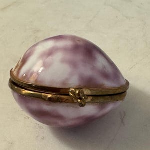 May include: A small, oval-shaped trinket box with a smooth, glossy finish featuring a marbled pattern of soft purple and white. The container is divided into two halves held together by a metallic gold-toned hinge and a small, decorative clasp.