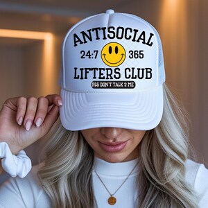 Puede incluir: Gorra de camionero blanca con el texto "ANTISOCIAL 24:7 365 LIFTERS CLUB PLS DON'T TALK 2 ME" en negro, una carita sonriente amarilla y una parte trasera de malla blanca.