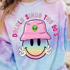Può includere: Una felpa tie-dye nei toni del rosa, viola e blu. La grafica presenta una faccina sorridente con segni di pace, un cappello a secchio rosa sfocato con un alieno e la scritta "Delulu Since The 90's."