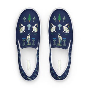May include: A pair of blue slip-on canvas shoes with a white sole. The shoes feature a repeating pattern of white rabbits, green trees, blue birds, and green crosses.